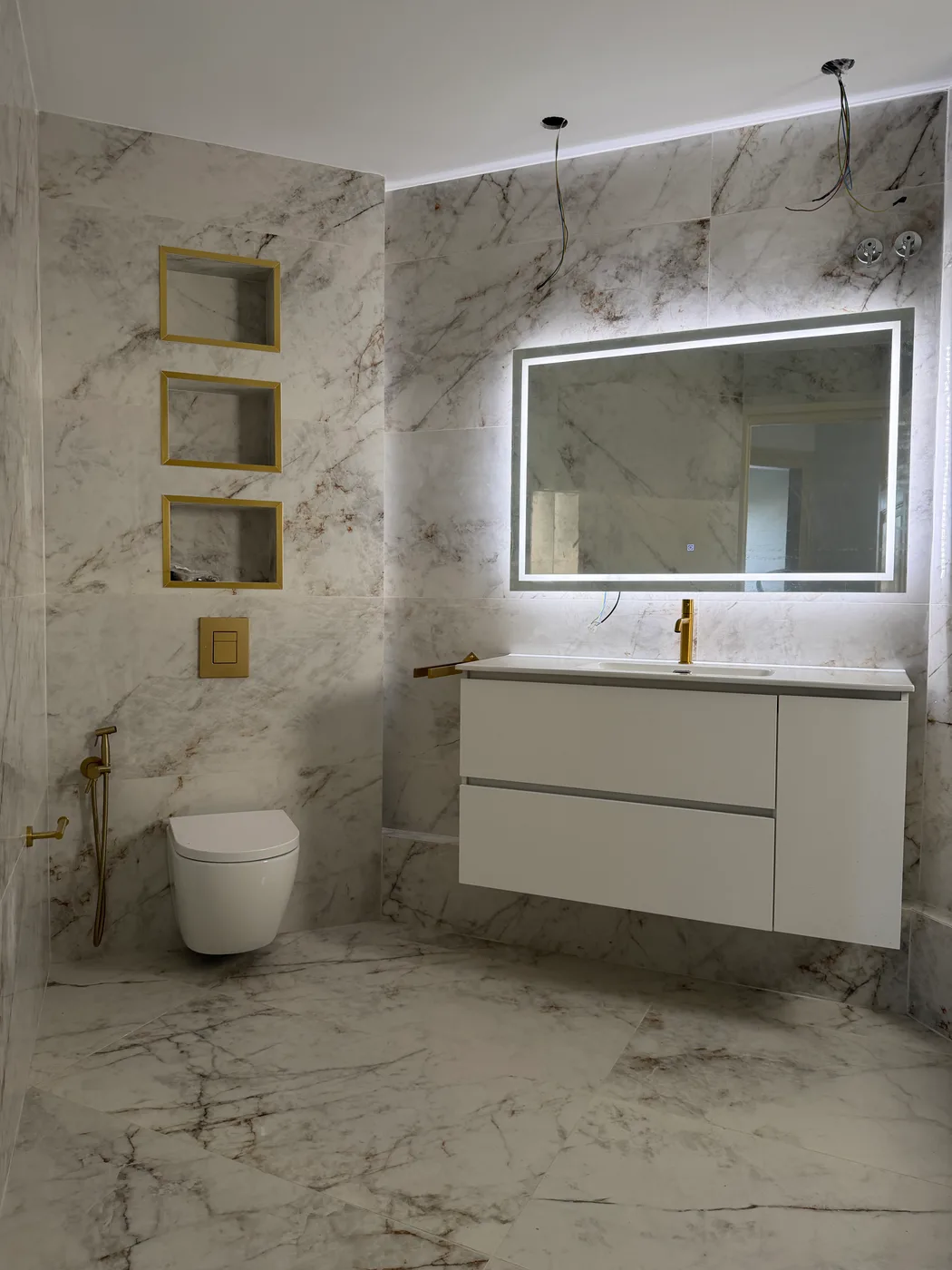 Baño revestido de cerámica efecto mármol, con espejo iluminado, mueble de lavabo suspendido y griferías doradas