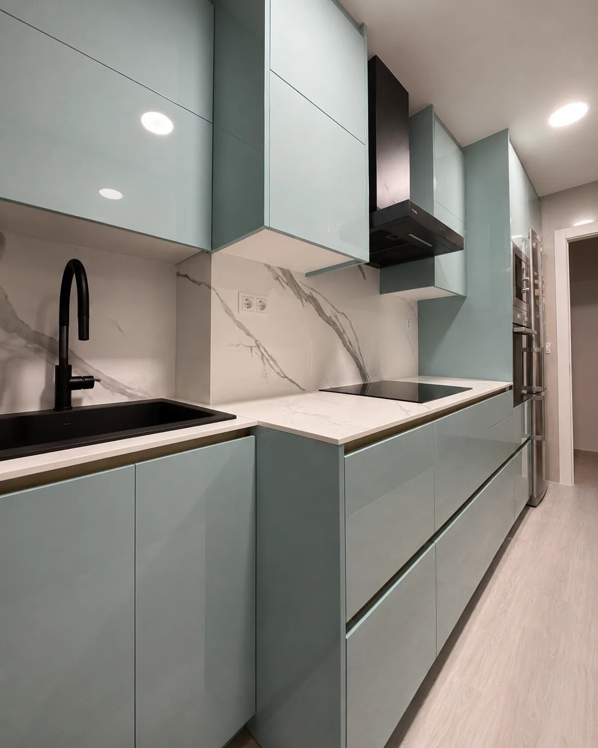 La misma cocina después de la reforma, con muebles verde agua hasta el techo, frontal porcelánico efecto mármol y fregadero negro