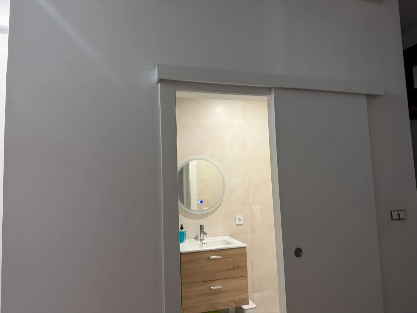 Baño acabado con mueble suspendido y espejo redondo tras una puerta corredera empotrada, reforma en Ciutat Vella, Barcelona