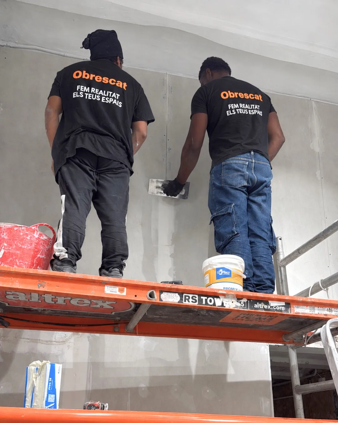 Dos operarios de Obrescat enlucen una pared subidos a un andamio