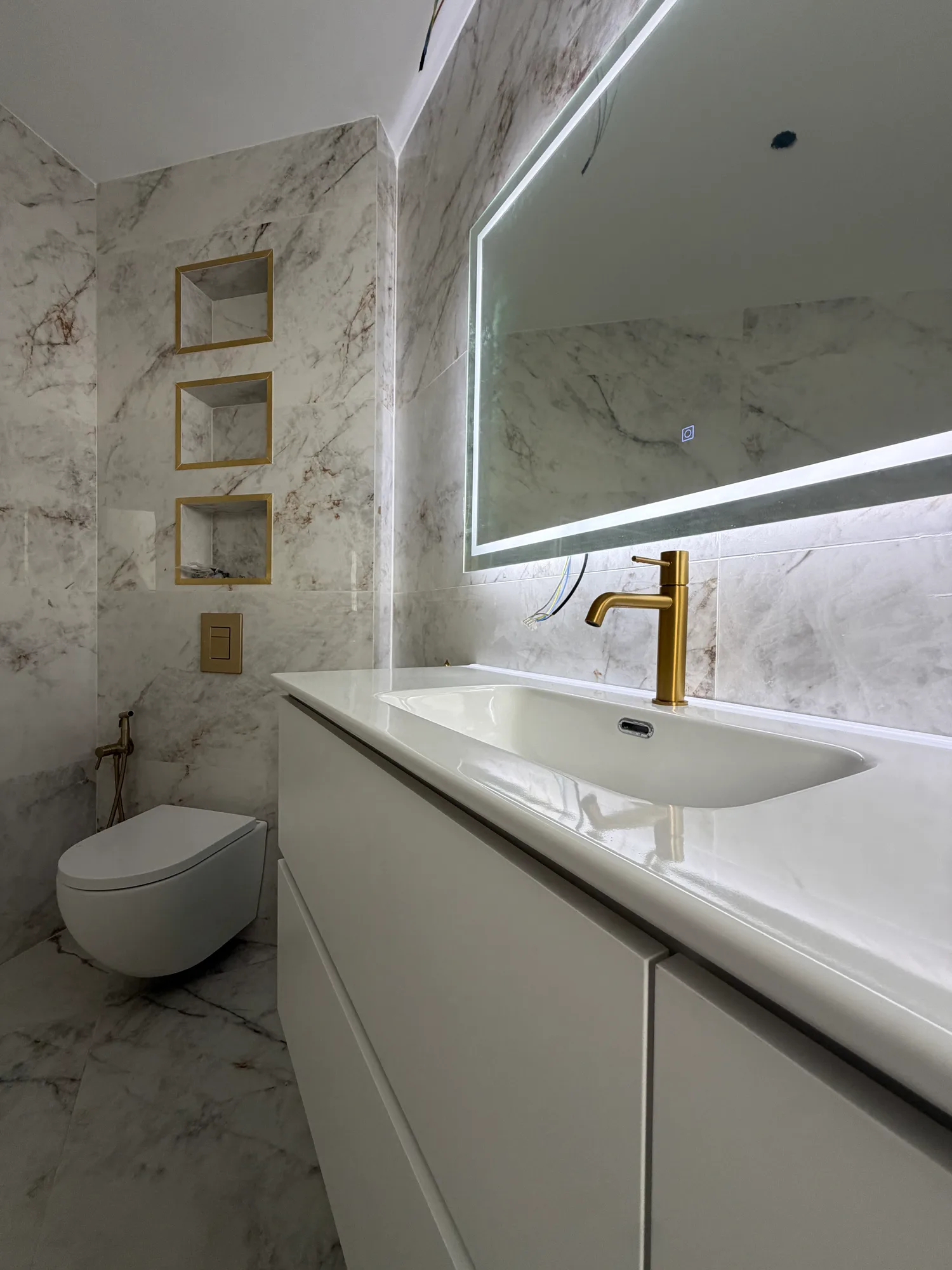Mueble de lavabo y espejo iluminado de un baño revestido de mármol, con grifería dorada y hornacinas junto al inodoro suspendido